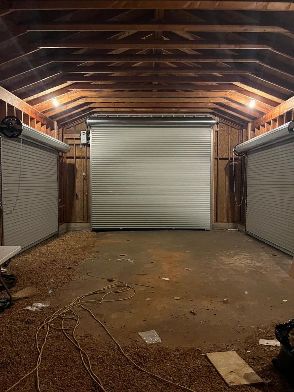Interior of a shed with roller doors and exposed wooden beams, showing dirt floor and stray wires.