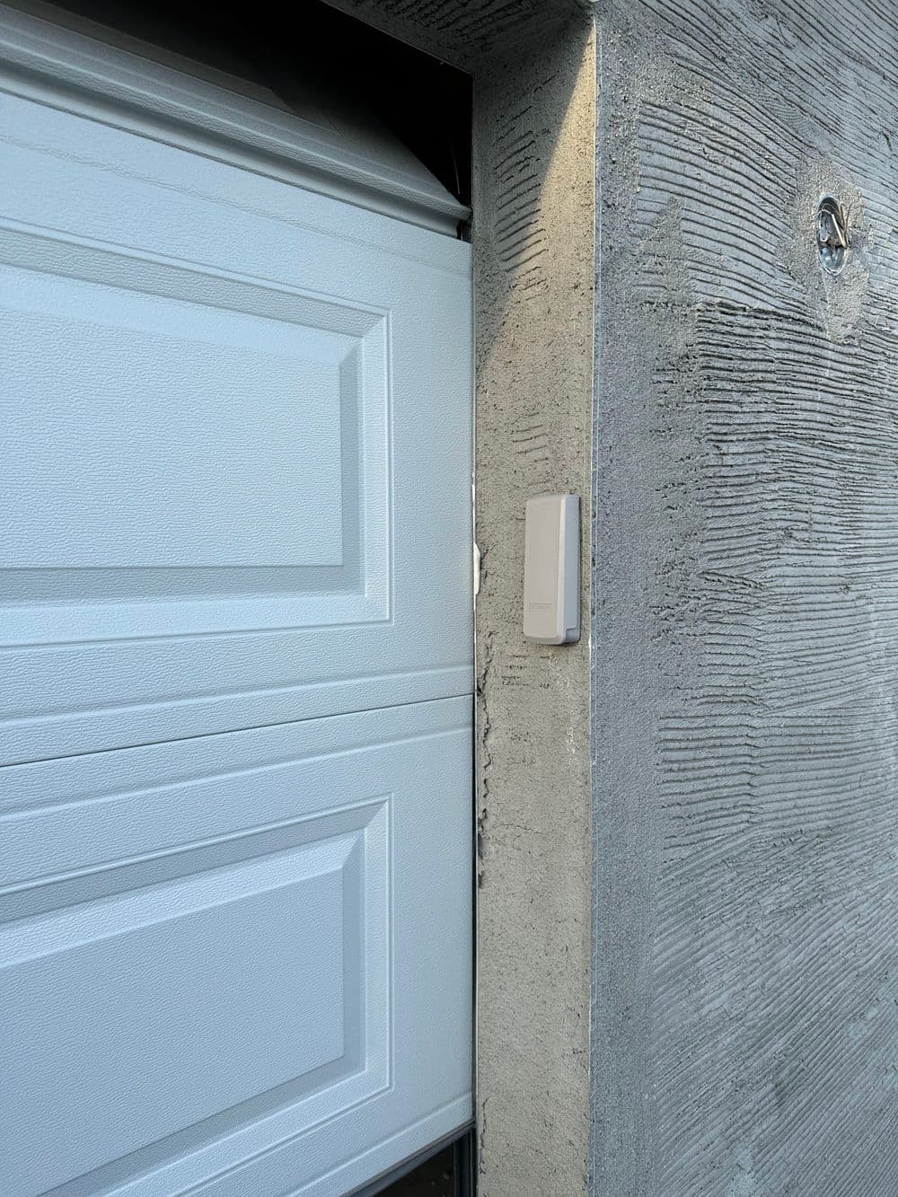 White garage door with a modern sensor mounted on a textured concrete wall.