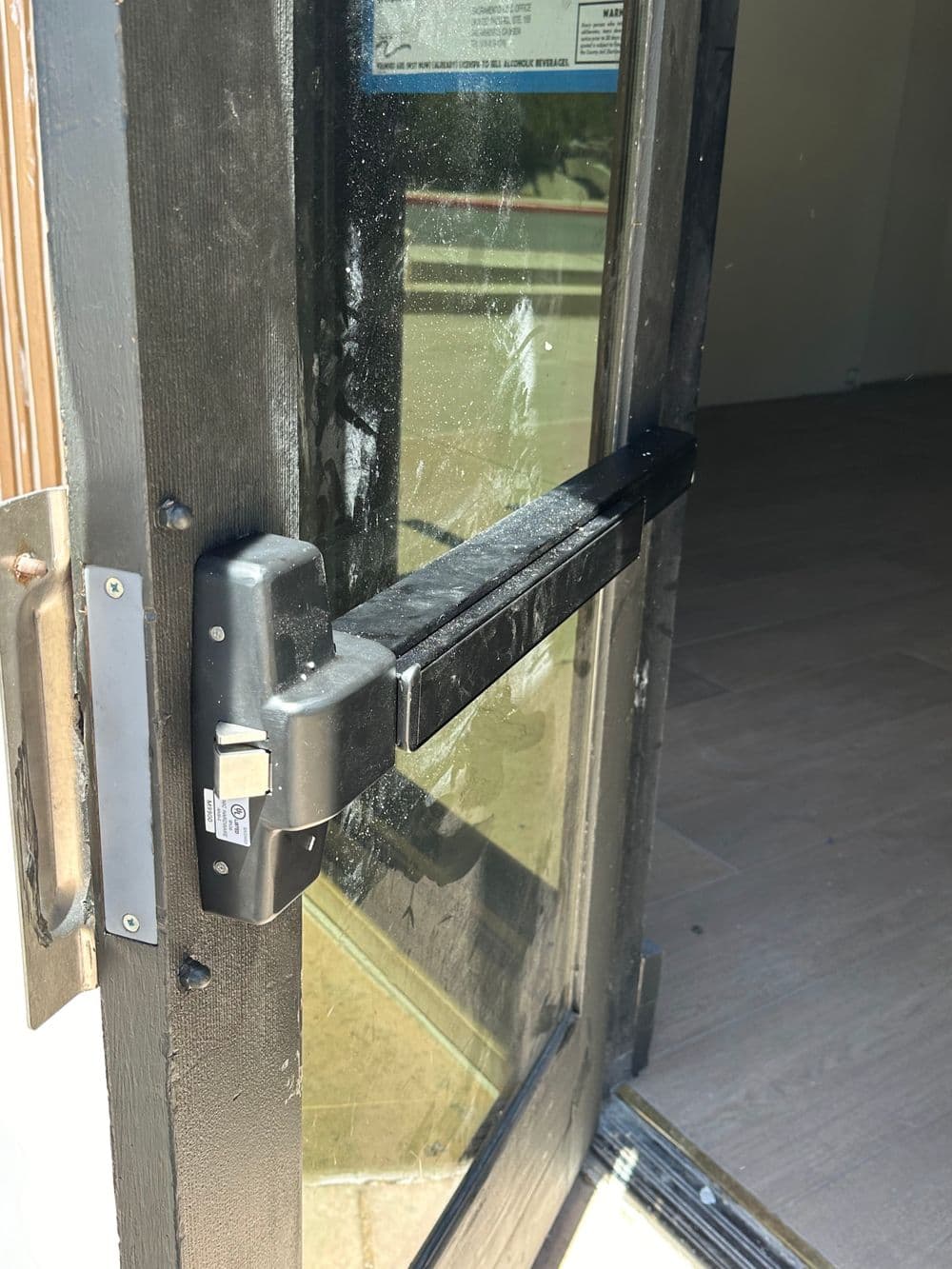 Black push bar door lock on a glass entry door with visible fingerprints and reflections.