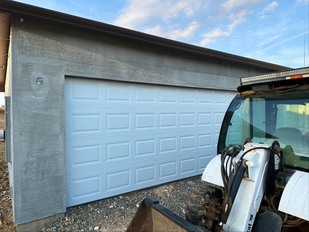 White garage door with textured concrete wall and construction vehicle on gravel surface.
