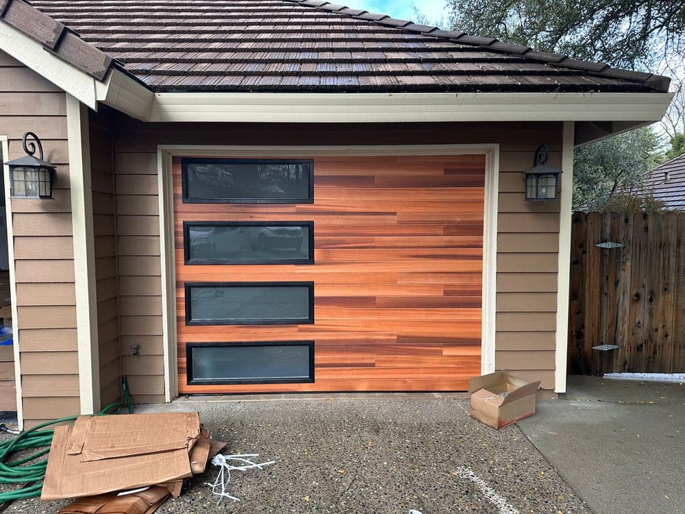 Modern wooden garage door with glass panels, surrounded by a rustic home exterior.