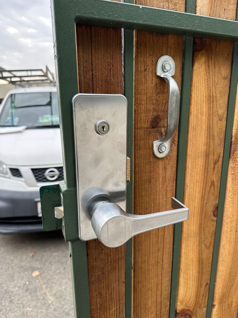 Modern gate lock on a wooden fence with a metal handle and a car in the background.