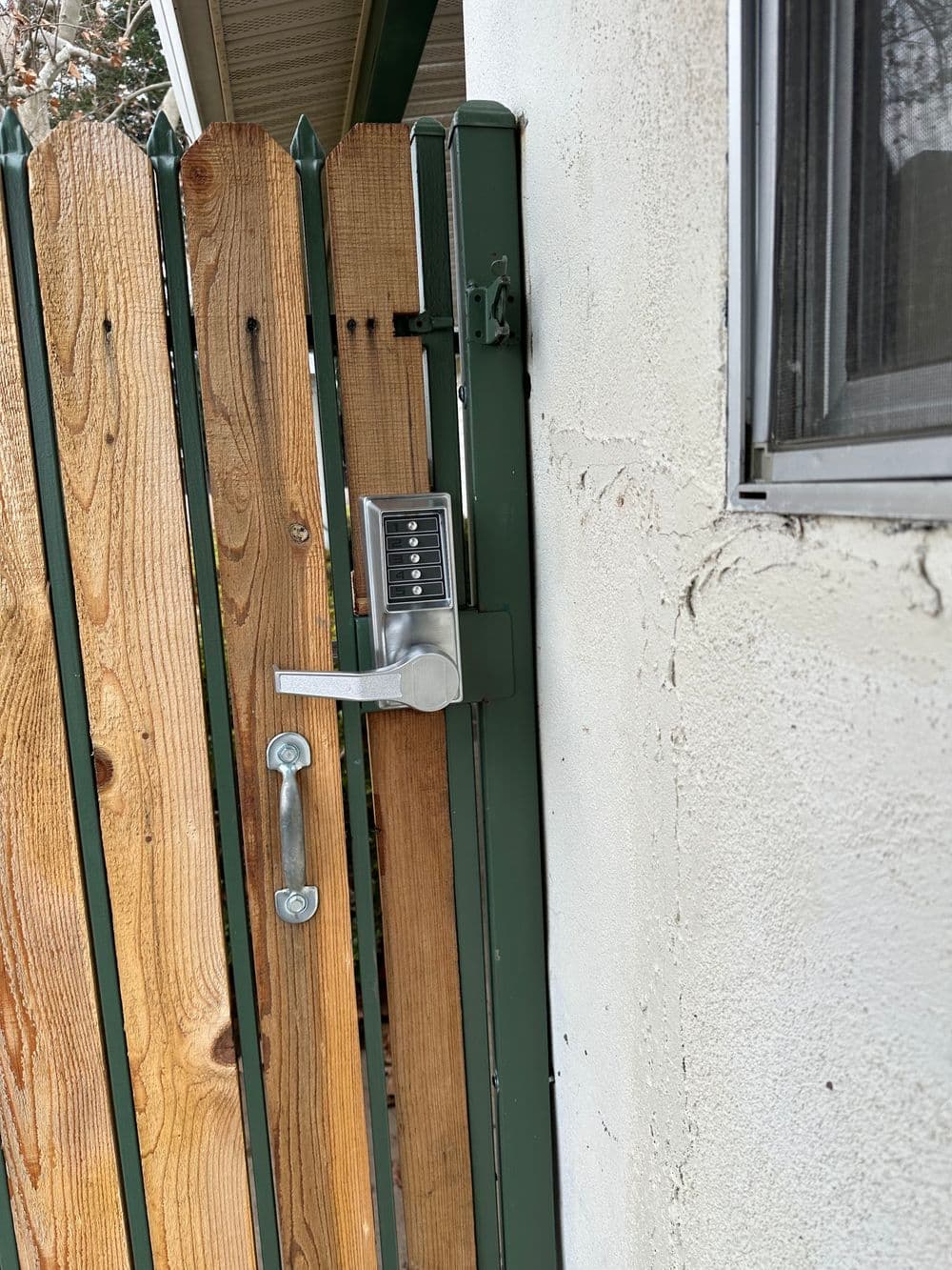 Keyless gate lock on wooden fence with green metal trim and adjacent wall.