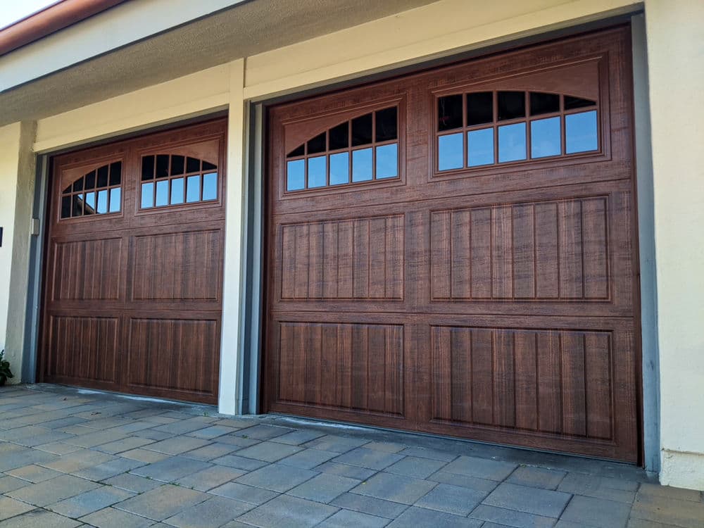 Modern wooden garage doors with arched windows and a textured finish on a stone driveway.