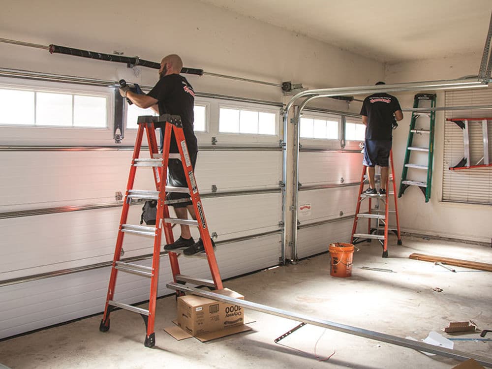 Two technicians installing a garage door system on ladders in an empty garage.