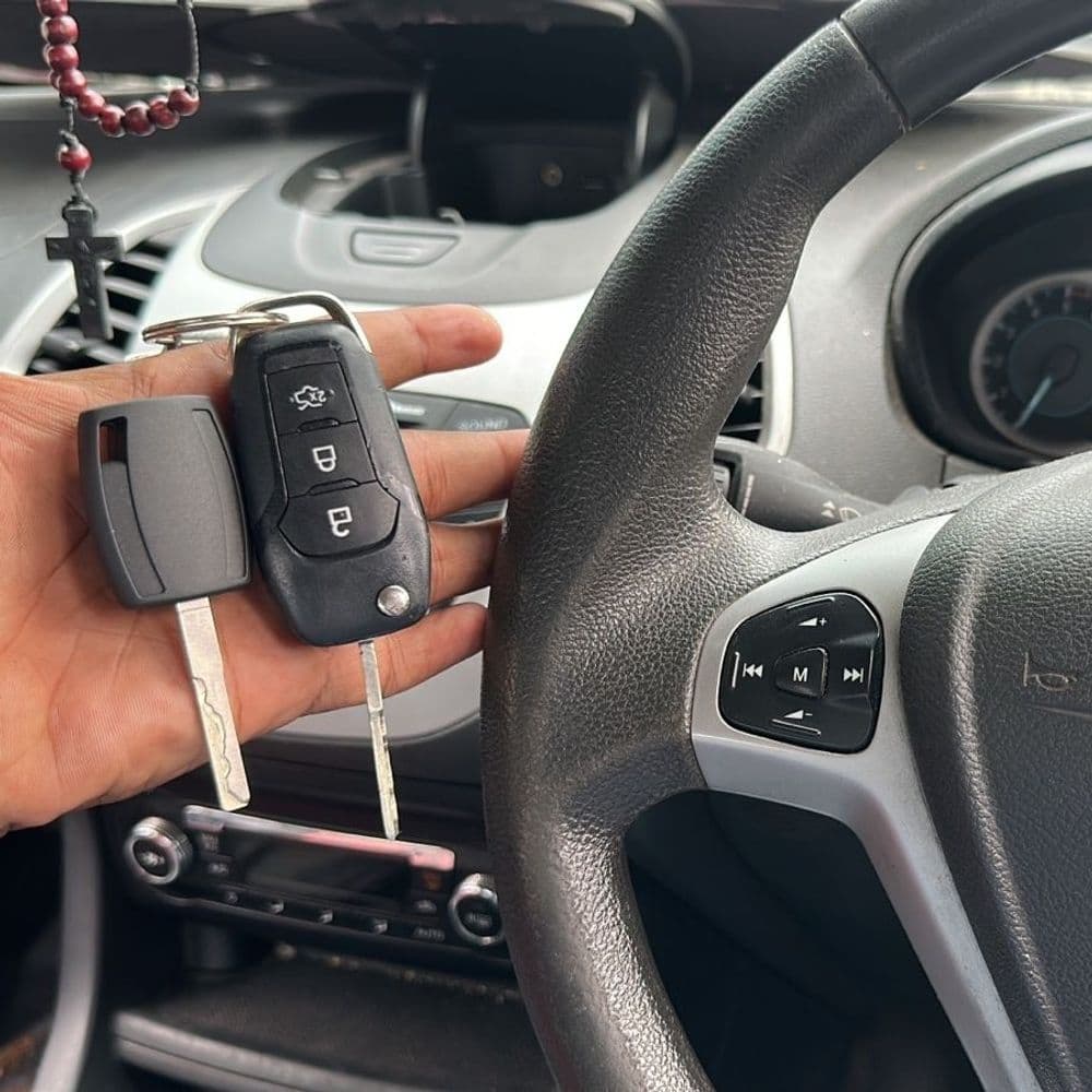 Hand holding car keys with a steering wheel and dashboard in the background.